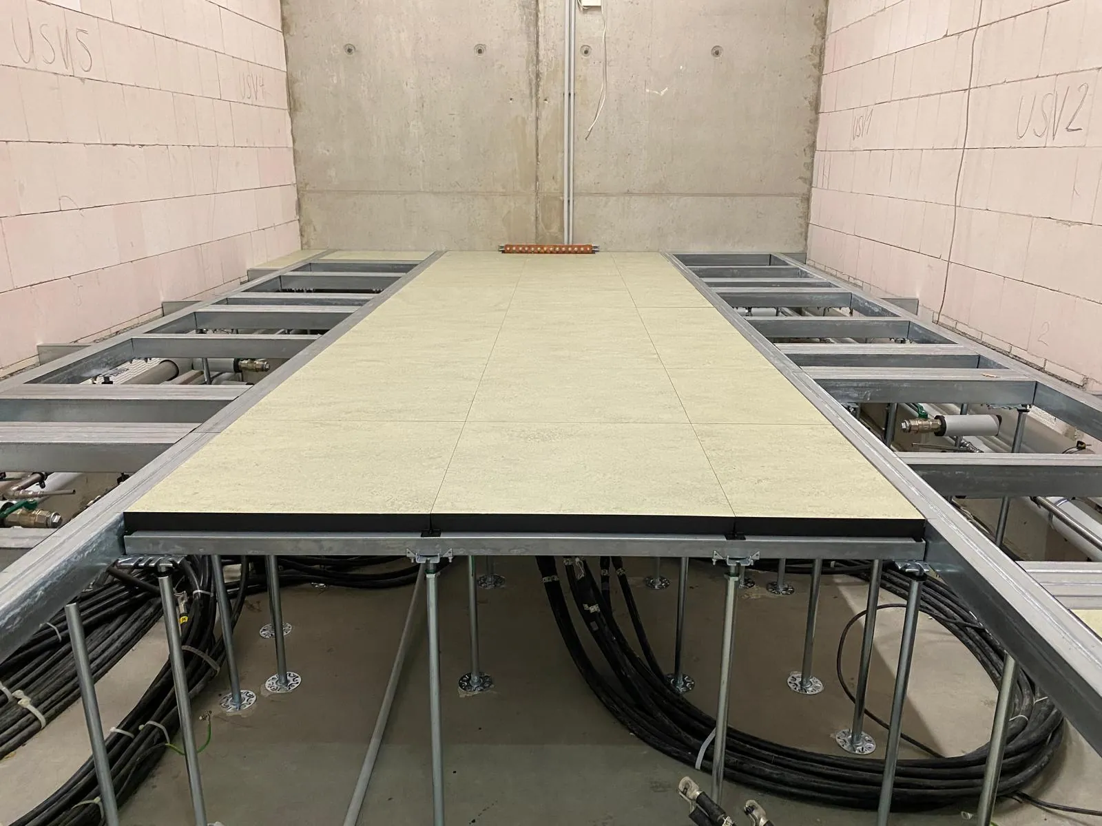 Doppelboden-Unterkonstruktion in einem Technikraum: Stützen, Rasterträger und Platten über Rohrleitungen und Kabeln